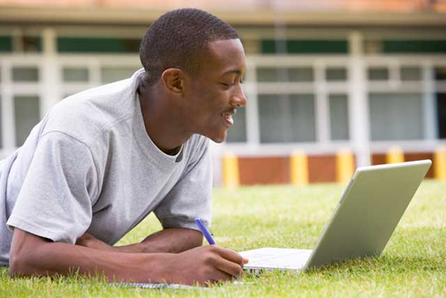 African-American male student using laptop on lawn while applying for college online.