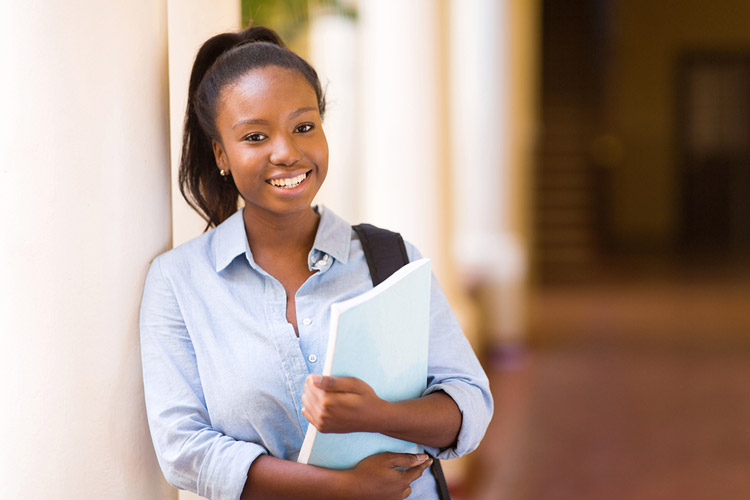 A Black female first generation college student poses on campus holding her books.