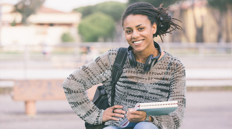 African-American female high school student sits outside with her books, headphones and backpack.