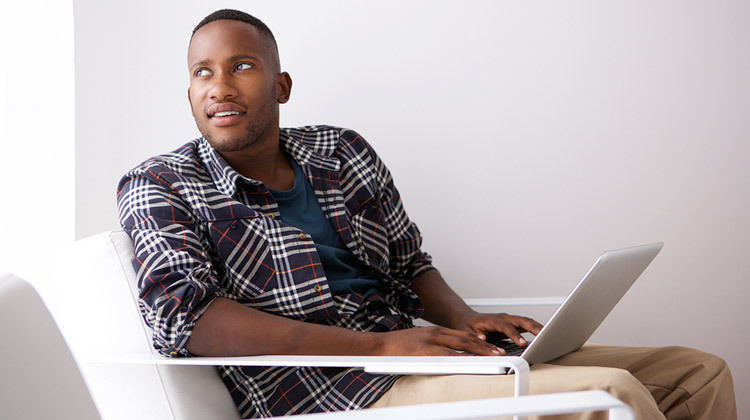 Before email your professor: A relaxed young black college student is sitting on a chair with a laptop, looking away and thinking of what to type.