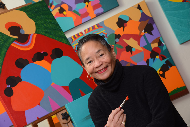 Renowned visual artist Synthia Saint James poses holding a brush with paint in front of her iconic paintings.